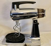 Side profile of vintage chrome Sunbeam Mixmaster mixer with black handle and control knob on black base plate, one beater attached