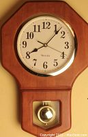 Full view of the wooden wall clock showing the octagonal frame, round clock face with black Arabic numerals, brass bezel, and brass pendulum visible in the lower glass panel.