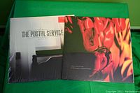 Two vinyl albums shown side by side on green background, The Postal Service 'Give Up' and Ulrich Schnauss 'A Strangely Isolated Place', both sealed in shrink wrap.