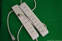 Two white WiFi enabled powerbars with six outlets each laid out on a green surface. Each powerbar has white cords with three-prong plugs.