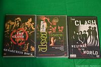 Three music DVDs placed side by side on a green surface, showing full front case covers of The Clash - Revolution Rock Live, The Doors - classic performances, and The Clash - Westway To The World.