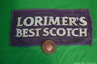 Photo of purple Lorimer's Best Scotch bar towel with white text and round metallic Levi Strauss belt buckle placed on green background.