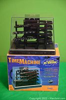 Front view of the Time Machine kinetic ball clock on original box, showing black frame with metal ball tracks and numeric indicators for time.