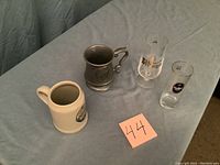 Four assorted beer mugs placed on a table with a numbered note '44'. Includes two clear glass mugs, one ceramic stein, and one pewter mug.