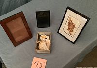 Lot showing two empty frames, a framed grape art piece, and a small angel figurine in a box on a blue cloth surface.