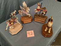 Four carousel horse music boxes shown on blue cloth background; includes Pegasus with wings, star horse, tiger, and dark brown horse with saddle; all on wooden bases