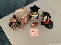 Photo showing side view of four ceramic animal items: a cat basket, an owl piggy bank with graduation cap and glasses, a small sitting cat, and a black dog with a red scarf. All items show wear.