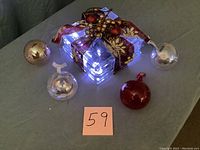 Four glass Christmas ornaments around a lit glass block tabletop decoration wrapped with burgundy and gold leaf ribbon.