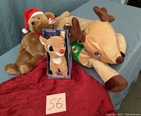Three Christmas stuffed animals on a blue surface: large plush reindeer, Ty Beanie Babies bear with Santa hat, singing Rudolph in original box, with red tablecloth beneath.