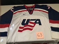Front view of USA hockey jersey showing USA logo with a star and flag, blue sleeves with red and white stripes, and a red-trimmed collar