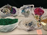 Six plastic bags containing Mardi Gras bead strands of various colors and styles including green, purple, white, gold, and pink.