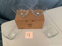 Four clear glass square dessert bowls arranged with two on a cardboard box and two in front of it. The bowls have an etched floral design on the sides.