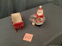 Santa figurine with clear dish top and small penguin attached to base, beside red ceramic sleigh with gold runners and holiday decorations on a gray surface.