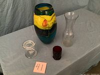 Four glass decorative items seen from slightly different angles showing each item's shape and color: a multicolored tall vase, a clear wine carafe, a small clear glass container with lid, and a deep red candle holder.