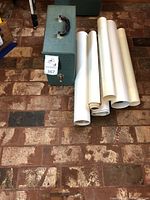 Green textured case with handle for a vintage projector shown next to several rolled maps or prints on brick flooring.