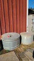 Two large round metal troughs side by side in outdoor setting with concrete or metal flat covers, one smaller at 36in diameter, one larger at 44in diameter.