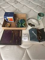 Full overview of lot contents on mattress including vintage drawing tools, mug, inflatable neck pillow, eyeglasses, picture frame, earthenware dish and cup, tape roll, beads, and keychain with magnifier.