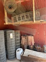 Overview of five rolls of fencing wire stored upright, assorted rolls of chicken wire and stacked tomato cages against red wall.