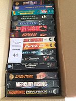 Top-down view of 15 VHS movie tapes in original cases inside a cardboard box showing titles like Iron Giant, Winnie the Pooh, The Matrix Reloaded, The Mask of Zorro, Speed, Mission Impossible, Pirates of the Caribbean.