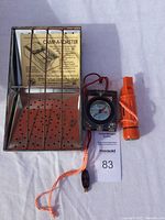 Top view of three camping items: Camp-A-Toaster with instructions inside, black compass, orange whistle with lanyard.
