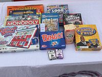 Lot of 11 assorted board games displayed on a white surface including Monopoly, Guess Who?, Family Feud, Yahtzee, Carcassonne, Bingo, Logo Party, and others.