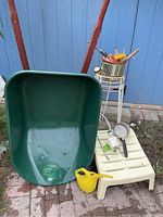 Photo showing green metal wheelbarrow with red frame, rotted wooden handles, yellow plastic watering can, small plastic footstool, metal plant stand, and various piping and garden tools on a patio.