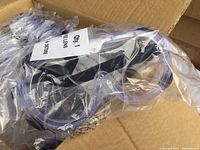 Photo showing close-up of transparent lens goggles with black frame, sealed in plastic wrapper with label.