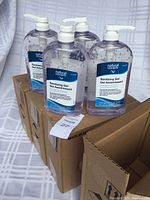 Four bottles of Natural Concepts Sanitizing Gel displayed on top of two open brown cardboard boxes. Bottles are clear plastic with white pump tops. Labels show product name and details.