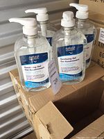 Front view of multiple bottles showing label and pump dispenser on top, arranged on top of two cardboard boxes.