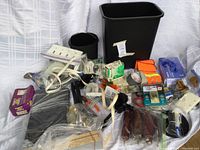 Various hardware and office items spread on a surface including power strips, cords, gloves, batteries, tape, small tools, and trash bins