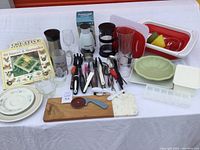 View of the entire kitchen lot on white surface, showing plates, glassware, cutting board, utensils, containers, and the chopper box.