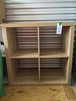 Full front view of a beige wood grain storage stand with four cubby compartments showing the overall shape and color.
