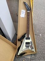 Full length image of the white Starfire Flying V style electric guitar with black pinstripes, in a cardboard box, showing headstock to body and attached black strap.