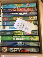 Box containing 10 children's VHS movies stacked showing spine titles including The Rescuers Down Under, Aladdin, Finding Nemo, Robin Hood, Spirit, Ice Age, The Fox and the Hound, Flubber, Fantasia 2000, and Spy Kids.