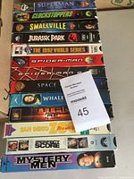 13 adult VHS movies stacked showing spines and titles such as Superman The Movie, Clockstoppers, Smallville, Jurassic Park, The 1992 World Series, Spider-Man, and Spider-Man 2.