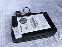 Sharp VC-H120 VHS player with instruction manual resting on top, showing overall unit and details of buttons and inputs.