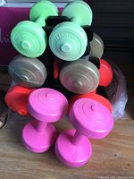 Colored dumbbell weights in light green, pink, orange, and grayish tan placed on a black plastic stand, showing front and top view