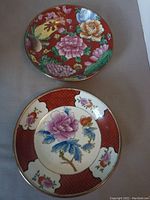 Two decorative Asian plates showing different colorful floral hand-painted designs with brass rims and hangers attached.