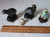 Four small cloisonné pieces displayed next to a ruler for size reference: one egg figure, two bird figures on wooden stands, and one thimble with owl design.