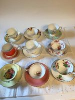 Full view of 9 vintage cup and saucer sets arranged in three rows on white background showcasing various floral and decorative designs