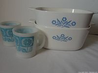 Front view showing two stacked white Corning casserole dishes with blue floral designs and two white Fire King mugs with blue geometric floral patterns.