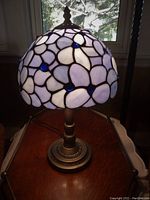Full frontal view of the blue stained glass lamp turned on showing flower petal pattern and blue glass jewels on shade, standing on wooden surface near window.