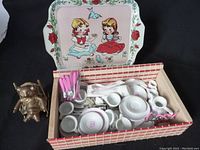 Box of miniature white china dishes and pink utensils alongside solid brass figurine and vintage illustrated tin tray in background