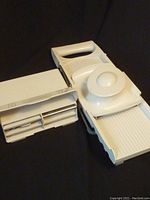 White plastic Pampered Chef mandolin slicer with handle, food holder with round grip, and blade storage container with four blades inside.
