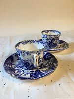 Two demitasse cups and saucers with blue and white Mikado pattern and gold trim, shown together on a cloth surface.