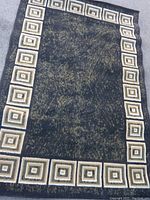 Full view of rectangular black area rug with beige square patterned border laid flat on ground