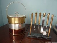 Ice bucket with tri-tone metal bands (gold, silver, copper), acrylic lid, and metal handle alongside brass and stainless steel bar tool set on green marble base