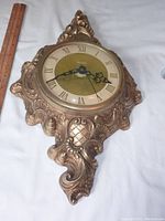 Full view of the vintage ornate United wall clock showing the round clock face with Roman numerals and decorative hands, set in an elaborate solid metal frame with intricate details and patterns. Clock is not working at the time of photographing.