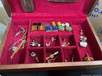 Wide view of the wooden jewelry box fully open showing contents of cuff links, tie clips, and other items arranged in individual compartments against red velvet lining.