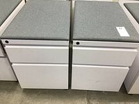 Two white metal file cabinets side by side with gray fabric cushioned tops and two drawers each, on wheels. Shows general condition and size.